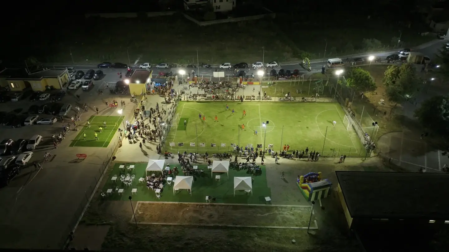 A floodlit five-a-side pitch at night seen from a drone, with a crowd along the fence, a volleyball court and white gazebos with tables beside it.